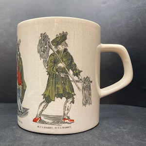 Könnte beinhalten: Eine weiße Keramiktasse mit craquelé-Glasur zeigt eine Vintage-Illustration einer Person, die ein Bündel Kaninchen trägt. Der Text "BUY A RABBET, BUY A RABBET" ist auf der Unterseite der Tasse aufgedruckt.