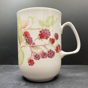 Może przedstawiać: Biały ceramiczny kubek z ręcznie malowanym wzorem jeżyn i liści. Owoce są czerwone i fioletowe, z zielonymi liśćmi i łodygami. Kubek ma zakrzywiony uchwyt. Wysokość około 10 cm.
