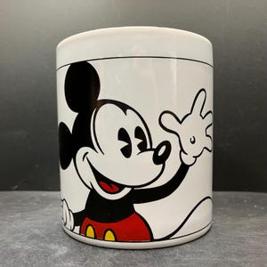 Puede incluir: Taza de cer&aacute;mica blanca con Mickey Mouse sonriendo y saludando. El personaje de dibujos animados est&aacute; representado en negro, rojo, amarillo y blanco. La taza tiene un contorno negro y un borde negro.