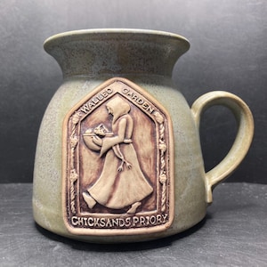 May include: A ceramic mug with a speckled, olive-green glaze. A rectangular, raised relief panel features a figure in a hooded robe, holding a basket, with the text "Walled Garden" and "Chicksands Priory". The mug has a rounded body and a curved handle.