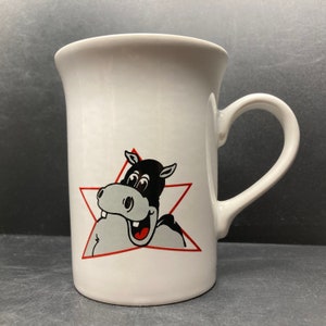 May include: White ceramic mug with a black and white cartoon hippopotamus illustration. The hippopotamus is smiling and has a red star behind it.
