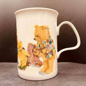 May include: A white ceramic mug featuring a colorful illustration of several teddy bears. One bear wears a multicolored checkered shirt, while another bear stands nearby wearing green shoes and holding a pink patterned blanket.