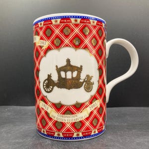 May include: A white ceramic mug with a red and gold pattern. The mug features a gold carriage design on a white background, with the text "Coronation HM Queen Elizabeth II" and the years 1953 and 2003.