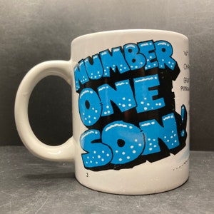 Peut inclure: Mug en céramique blanche avec l'inscription "NUMBER ONE SON!" en bleu et noir, dans un style de dessin animé audacieux. La tasse a une anse incurvée et le chiffre "3" imprimé en bas.