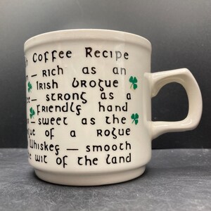 Op de afbeelding: Crèmekleurige keramische mok met een handvat. De mok heeft zwarte tekst en groene klaveraccenten. De tekst luidt "Coffee Recipe" gevolgd door beschrijvende zinnen. Een charmante en unieke koffiemok.