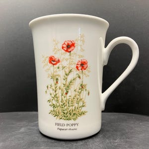 Könnte beinhalten: Weißer Keramikbecher mit einem Feldmohn-Design mit roten Blüten und grünem Laub. Der Becher hat einen gebogenen Henkel und die Worte "FIELD POPPY Papaver rhoeas" sind auf der Vorderseite aufgedruckt.