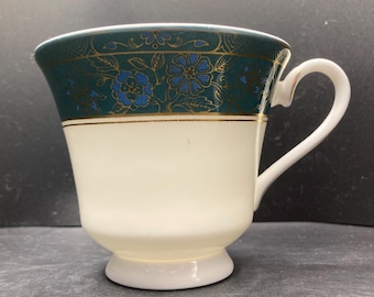 May include: A white teacup with a teal and gold floral pattern around the rim. The cup has a white handle and sits on a small, white pedestal base. The floral design includes blue flowers.