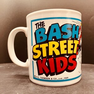 May include: A white ceramic mug featuring the colourful text THE BASH STREET KIDS in blue, yellow, and red. The design includes a partial cartoon illustration on the right side and the copyright text D.C. THOMSON & CO., Ltd., 1991 at the bottom.