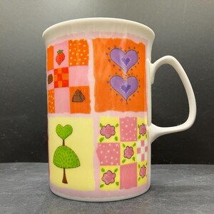 Könnte beinhalten: Weiße Keramiktasse mit einem skurrilen Design. Die Tasse hat ein Patchworkmuster mit orangefarbenen, rosa- und gelbfarbenen Quadraten, die mit Herzen, Erdbeeren und Blumen verziert sind. Der Griff ist weiß und gebogen.