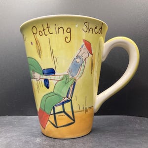 以下が含まれることがあります： 「Potting Shed」の文字と、椅子に座ってくつろぐ男性の気まぐれなイラストが描かれた黄色のセラミックマグ。 マグには白いハンドルと黄色の内側があります。