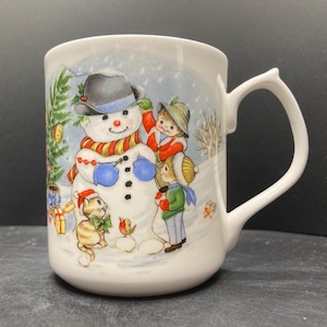 May include: White ceramic mug with a whimsical winter scene. The mug features a snowman with a black hat and a colorful scarf, children, a cat, and a bird. The handle has a unique curved design. The mug is decorated with a Christmas tree and gifts.