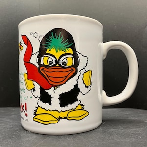 May include: White ceramic mug featuring a cartoon duck wearing a black aviator mask, a red scarf, and a black jacket with white trim. The duck has yellow feet and a green mohawk. The mug has a white handle and text on the side.