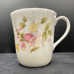 May include: White ceramic mug with a floral design featuring white flowers, pink buds, and green leaves. The mug has a curved handle and a slightly flared rim. The floral pattern is delicate and detailed, adding a touch of elegance to the mug.