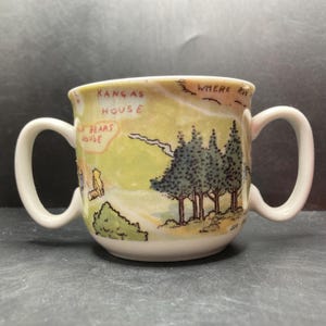 May include: A white ceramic mug with two handles features a Winnie the Pooh map design. The map shows the locations of "Kanga's House", "Piglet's House", and "Where Roo is".