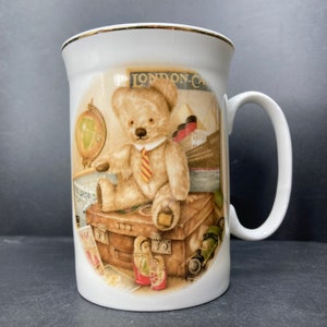 May include: White ceramic mug with a gold rim featuring a teddy bear wearing a tie sitting on a suitcase with a globe and other travel items. The mug has the text "LONDON-CAL" on it.