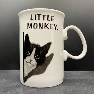 Peut inclure: Mug en céramique blanche avec une illustration en noir et blanc d'un chat regardant par-dessus un mur. Le mug porte l'inscription "LITTLE MONKEY." en noir. Il possède une anse incurvée.