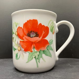 May include: White ceramic mug decorated with a large red poppy flower, dark center, and green leaves. The mug also features white daisies and green buds. The handle is white and curved. The mug is suitable for tea or coffee.