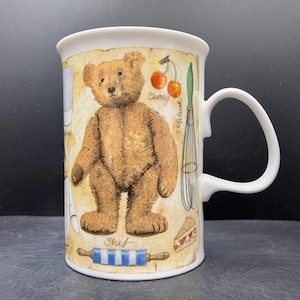 May include: A white ceramic mug with a brown teddy bear design. The mug has a handle and is decorated with baking-related images, including cherries, a whisk, and a rolling pin. The word "Chef" is also visible.