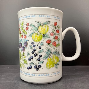 Könnte beinhalten: Weiße Keramiktasse mit farbenfrohem Fruchtdesign. Die Tasse zeigt Illustrationen von Kirschen, Äpfeln, Birnen, Pflaumen und Beeren. Der Rand der Tasse trägt die Worte "CHERRIES • APPLES • PEARS" in blauer Schrift.