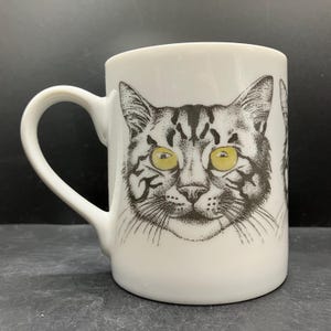 May include: White ceramic mug with a detailed illustration of a cat's face. The cat has yellow eyes and a black and grey patterned coat. The mug has a curved handle and is suitable for hot or cold drinks.