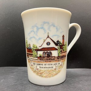May include: White ceramic mug featuring a detailed illustration of a church with a red roof, surrounded by trees and a brick wall. The mug has a curved handle and the text "THE SHRINE OF OUR LADY OF WALSINGHAM" on a banner.
