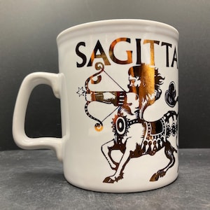 Op de afbeelding: Witte keramische mok met een gouden Boogschutter sterrenbeeld ontwerp. De mok heeft het woord "SAGITTA" in gouden letters boven een boogschutter met pijl en boog. Het handvat is wit.