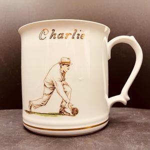May include: A white ceramic mug featuring a gold-coloured name Charlie printed near the top rim. The mug displays a detailed illustration of a person in a hat and light-coloured clothing in a bowling stance, holding a ball on a small patch of green.