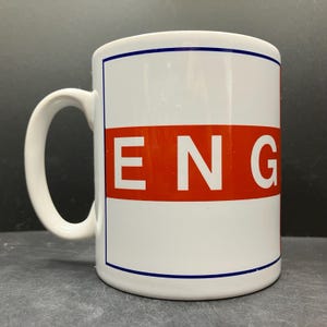 May include: White ceramic mug with a blue border and handle. The mug features a wide orange stripe with the letters "ENG" in white. The mug is designed for hot beverages.