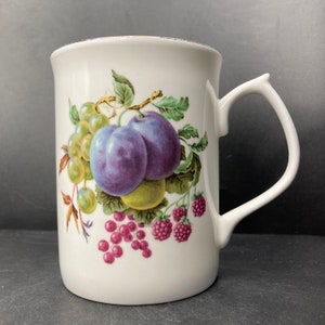 Könnte beinhalten: Weiße Keramiktasse mit einem bunten Fruchtdesign. Das Design zeigt Pflaumen, Trauben, Himbeeren und Johannisbeeren.