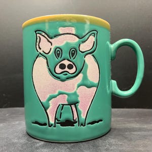 Könnte beinhalten: Eine türkisfarbene Keramiktasse mit einer skurrilen Schweine-Illustration. Das Schwein ist rosa mit schwarzen Umrissen und ist auf der Tasse zentriert. Der Rand ist gelb und der Henkel türkis. Eine dekorative Tasse.