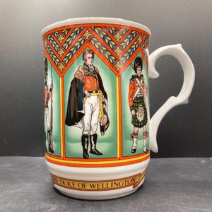 May include: White ceramic mug featuring illustrations of historical figures in military attire. The mug has a decorative handle and a band at the base with the text "THE DUKE OF WELLINGTON". The mug's design includes red, orange, green, and blue colors.