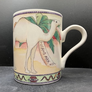 Könnte beinhalten: Cremefarbene Keramiktasse mit einer Kamelillustration und dem Text "NOAH'S ARK". Die Tasse hat einen gebogenen Henkel und einen dekorativen Rand mit einem sich wiederholenden Muster aus Häusern und Schlangen. Das Kamel ist vor Palmen platziert.