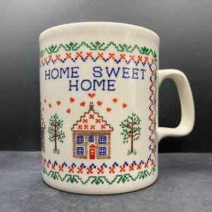 May include: Cream ceramic mug with a handle, decorated with a cross-stitch pattern. The mug displays the words "HOME SWEET HOME" in blue, along with images of houses and trees in red, green, and brown.