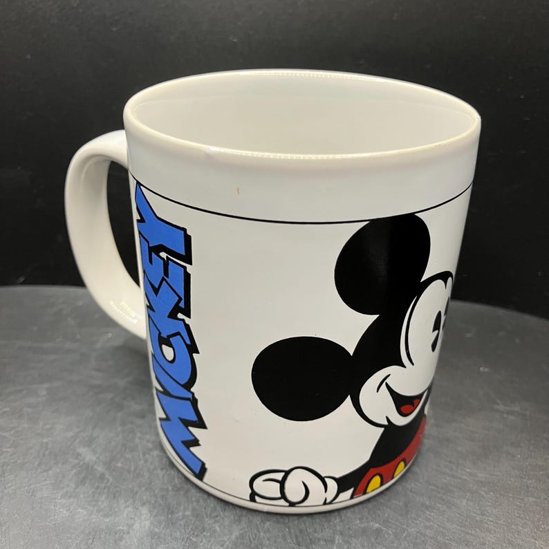 Puede incluir: Taza de cer&aacute;mica blanca con un dibujo de Mickey Mouse sonriendo. La taza presenta un dise&ntilde;o negro y rojo de Mickey Mouse en un lado y la palabra "MICKEY" en azul en el otro. Tiene un asa blanca y un fondo negro.