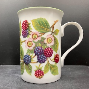 May include: White ceramic mug with a detailed botanical design featuring blackberries, pink flowers, and green leaves. The mug has a white handle and a green rim. The design wraps around the mug.