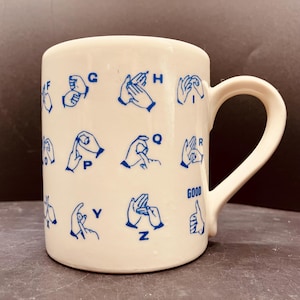 May include: A cream-coloured ceramic mug featuring blue printed illustrations of American Sign Language hand signs for letters F through Z, along with the word GOOD and a thumbs-up gesture. The mug has a curved handle on the right side.