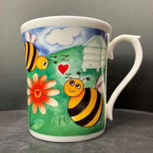 May include: White ceramic mug with a whimsical bee-themed design. The mug features cartoon bees, a red heart, a daisy, and a beehive against a green and blue background. The handle is white and has a unique shape.