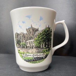 May include: White ceramic mug with a black and white illustration of Ripon Cathedral, a historic building with a tall tower and a green tree in front of it. The text "Ripon Cathedral" is written in a decorative font on the mug.