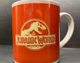 May include: A red ceramic mug with a white handle. The mug features a yellow Jurassic World logo with a dinosaur silhouette.
