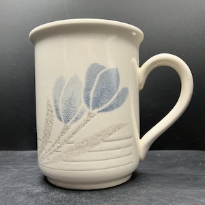 May include: Cream ceramic mug with a blue floral pattern. The mug has a curved handle and a cylindrical shape. The floral design features blue flowers and grey leaves. Suitable for hot or cold drinks. Capacity approximately 300 ml.
