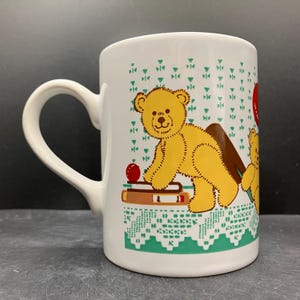 Könnte beinhalten: Weiße Keramiktasse mit einem gelben Teddybären-Design. Die Tasse hat einen gebogenen Henkel und ist mit einem sich wiederholenden Muster aus grünen Dreiecken und einem grünen spitzenartigen Rand verziert. Der Teddybär ist mit einem roten Apfel und Büchern dargestellt.
