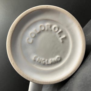 Puede incluir: Primer plano de la base de una taza de cer&aacute;mica blanca. La base est&aacute; ligeramente empotrada con el texto en relieve "COLOROLL ENGLAND" en un patr&oacute;n circular. El borde de la taza es de color crema.