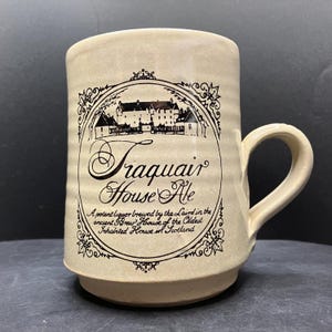 May include: A beige ceramic mug with a handle. The mug features a black and white illustration of a building and text that reads "Traquair House Ale". The text also mentions the ancient Brew House of the oldest inhabited house in Scotland.