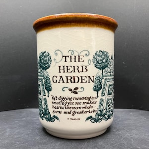 May include: A ceramic herb garden container with a brown rim and a white body. The container features a teal garden design with the words "THE HERB GARDEN" and a quote by T. Tusser.