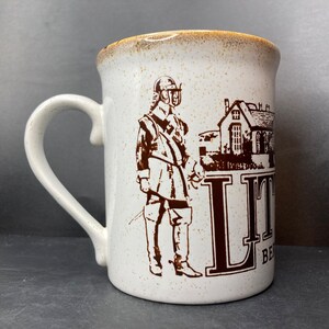 May include: White ceramic coffee mug with a brown illustration of a man in a helmet and a building with the text "Little Bedford" on the side.