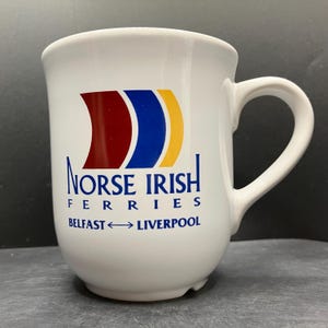 Op de afbeelding: Witte keramische mok met een gebogen handvat. De mok heeft de tekst "NORSE IRISH FERRIES" en de route "BELFAST ↔ LIVERPOOL" in blauw. Het logo bevat rode, blauwe en gele gebogen vormen.