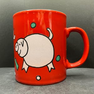 Könnte beinhalten: Eine rote Keramiktasse mit einer Cartoon-Schwein-Illustration. Das Schwein ist weiß mit schwarzen Umrissen und rosa Füßen. Grüne und weiße dekorative Elemente sind um das Schwein herum verstreut. Die Tasse hat einen abgerundeten Griff.