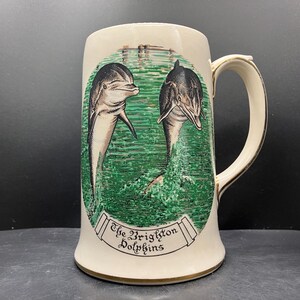 Może przedstawiać: Kremowy ceramiczny kufel do piwa ze złotym uchwytem i podstawą. Kufel przedstawia dwa delfiny wyskakujące z zielonej wody, z napisem "The Brighton Dolphins" na bannerze poniżej.