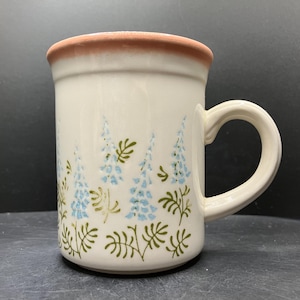 May include: Cream ceramic mug with a brown rim and a curved handle. The mug features a floral pattern of blue and green stylized flowers and foliage. The mug is cylindrical in shape.