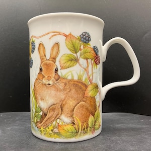 May include: White ceramic mug featuring a detailed illustration of a brown rabbit amidst foliage and blackberries. The mug has a curved handle and a glossy finish, perfect for enjoying hot beverages. The design evokes a woodland theme.